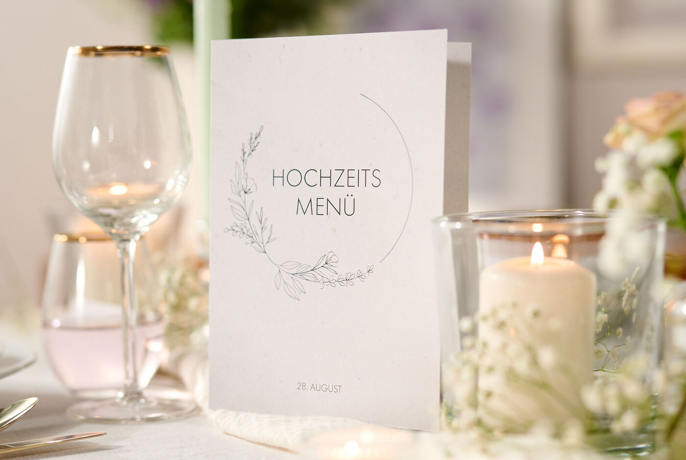 Auf einem mit Gläsern, Blumen und Kerzen gedeckten Tisch steht eine Klappkarte mit floralem Design und dem Titel "Hochzeitsmenü"