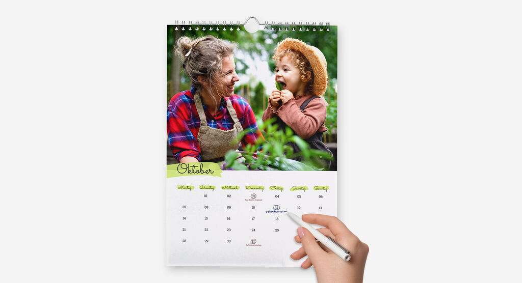 Eine Hand markiert mit einem Kugelschreiber einen Geburtstag im Kalender, der ein Foto von Frau mit Kind im Garten zeigt.