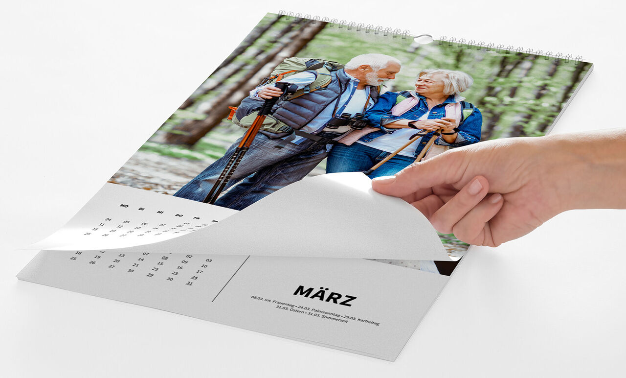 Eine Hand blättert von einem Kalenderblatt mit einem Foto eines älteren Paares beim Wandern zum nächsten Monat.