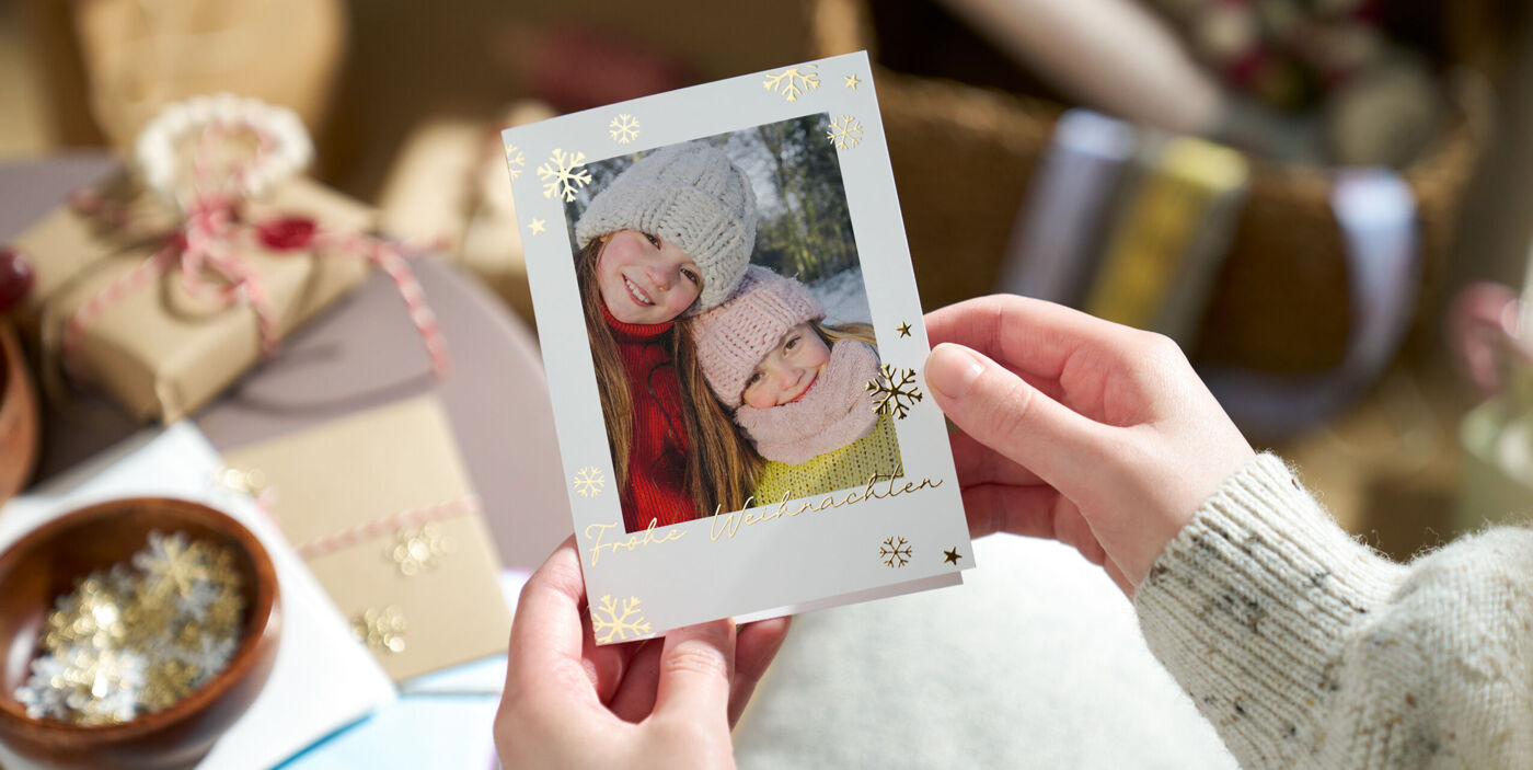Hände halten eine weihnachtliche Grußkarte vor sich, die mit goldenen Schneeflocken und einem Kinderfoto bedruckt ist
