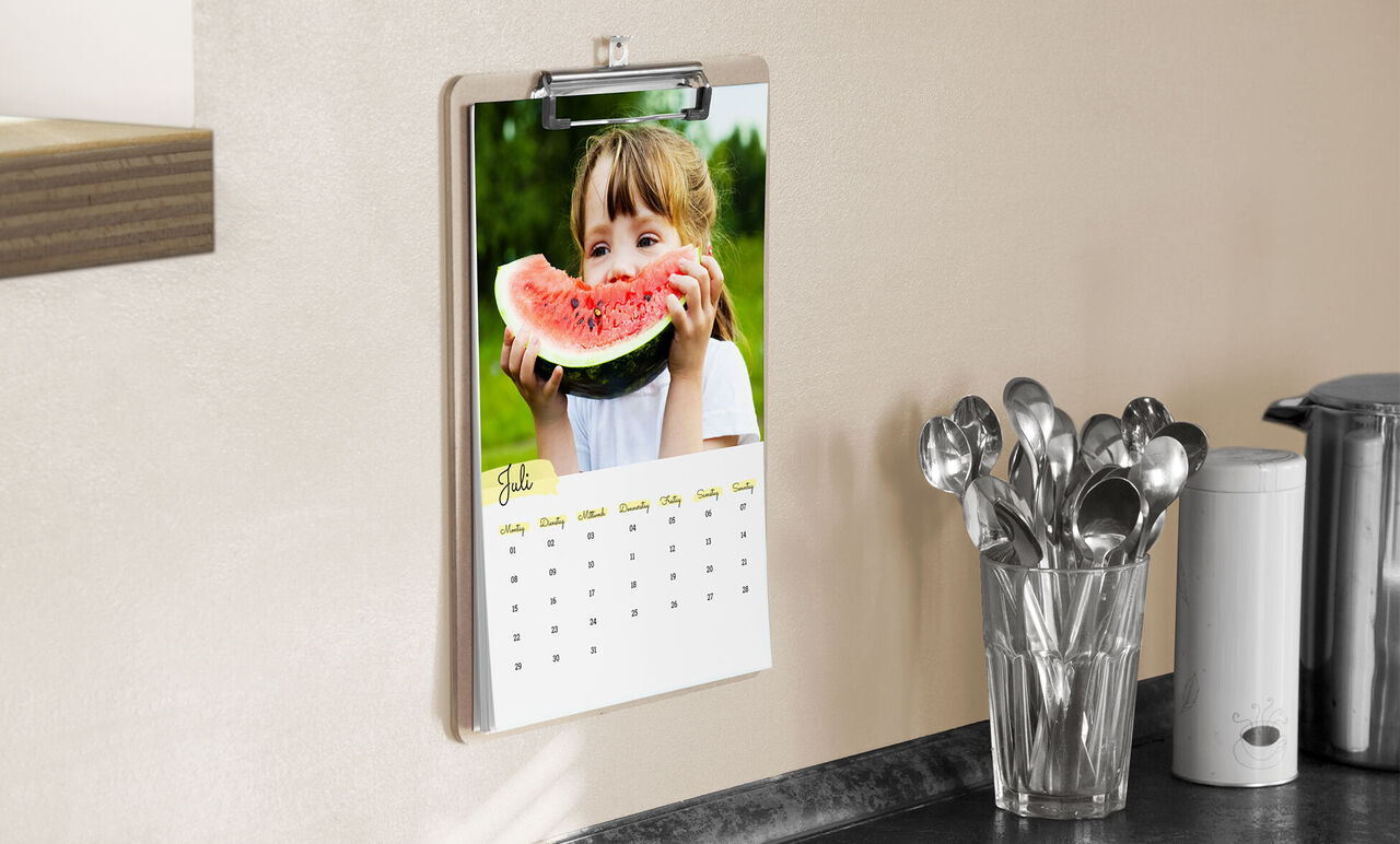 Kalender mit Klemmbrett, Design mit Farbakzenten und Foto von Mädchen mit Melonenstück, hängt an der Wand in der Küche.