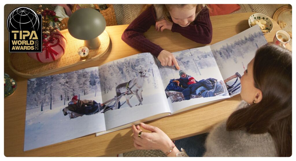 Eine Familie betrachtet ein CEWE FOTOBUCH mit Panoramaseite auf einem Tisch. Die Seite zeigt ein Foto von Kindern auf Rentierschlitten