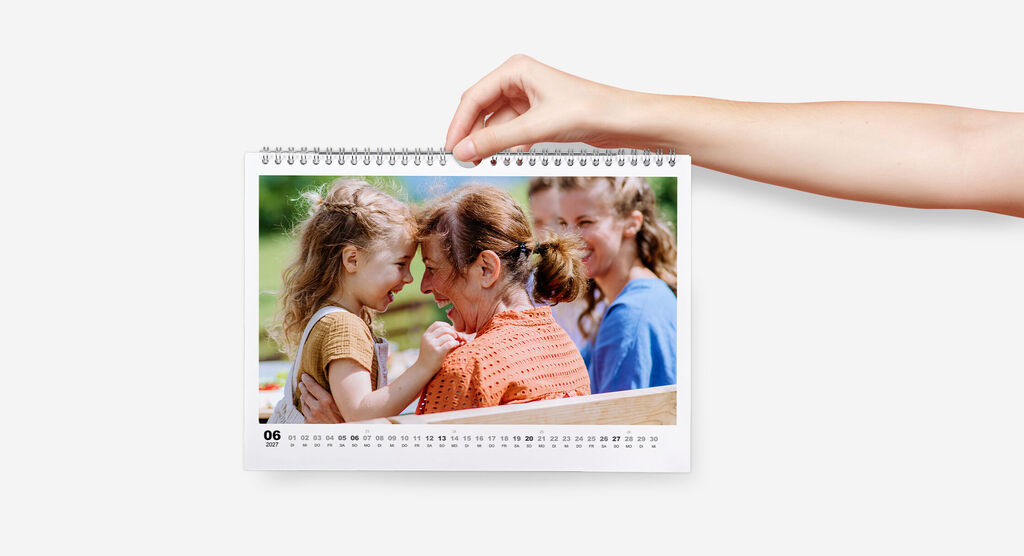 Wandkalender im Querformat mit Spiralbindung mit weißem Design und Foto von Oma mit Enkelin wird von einer Hand gehalten.