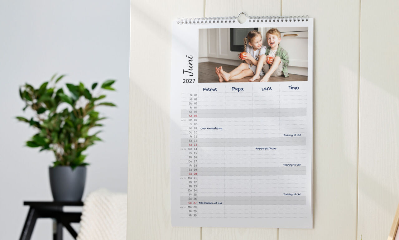 Familienkalender mit Spiralbindung, Kinderfoto und einem Tabellendesign mit Spalten für vier Personen, hängt an der Wand.