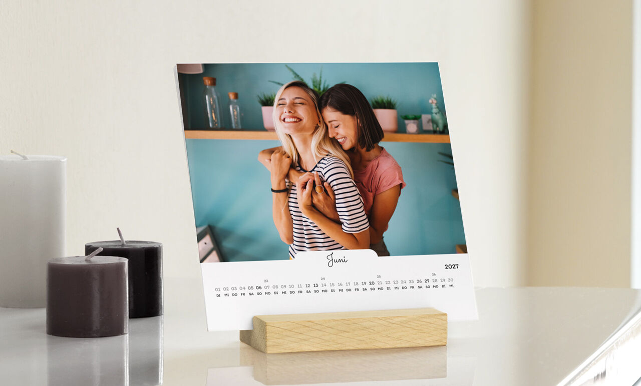 Quadratischer Tischkalender im Format 15 x 15 cm im Holzaufsteller. Das vordere Blatt zeigt ein Foto von zwei Frauen, die sich umarmen
