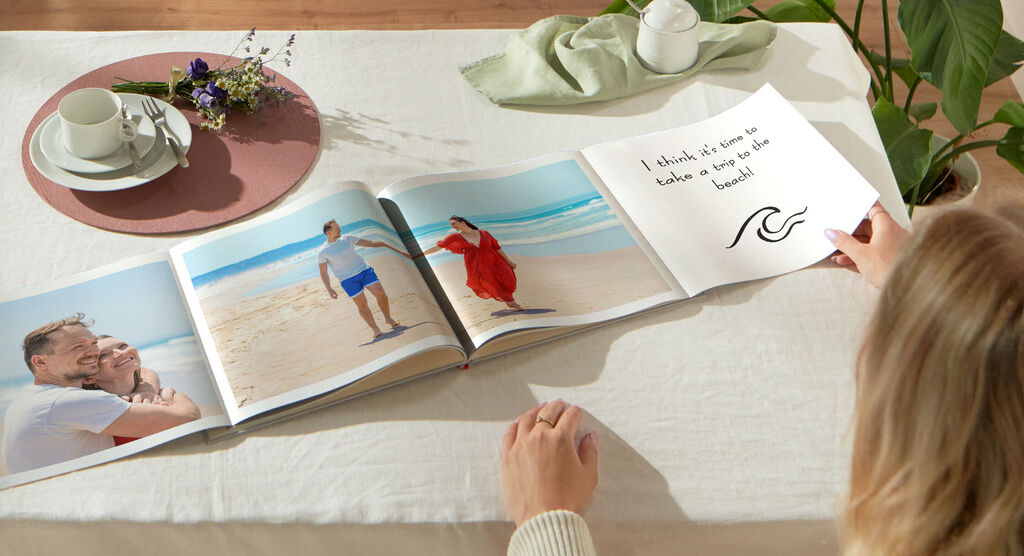 Ein offenes Fotobuch auf einem Tisch. Eine Person blättert durch die Seiten mit schönen Urlaubsfotos am Strand.