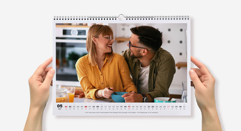 Wandkalender im Querformat mit Spiralbindung mit weißem Design und Pärchenfoto wird von zwei Händen seitlich gehalten.