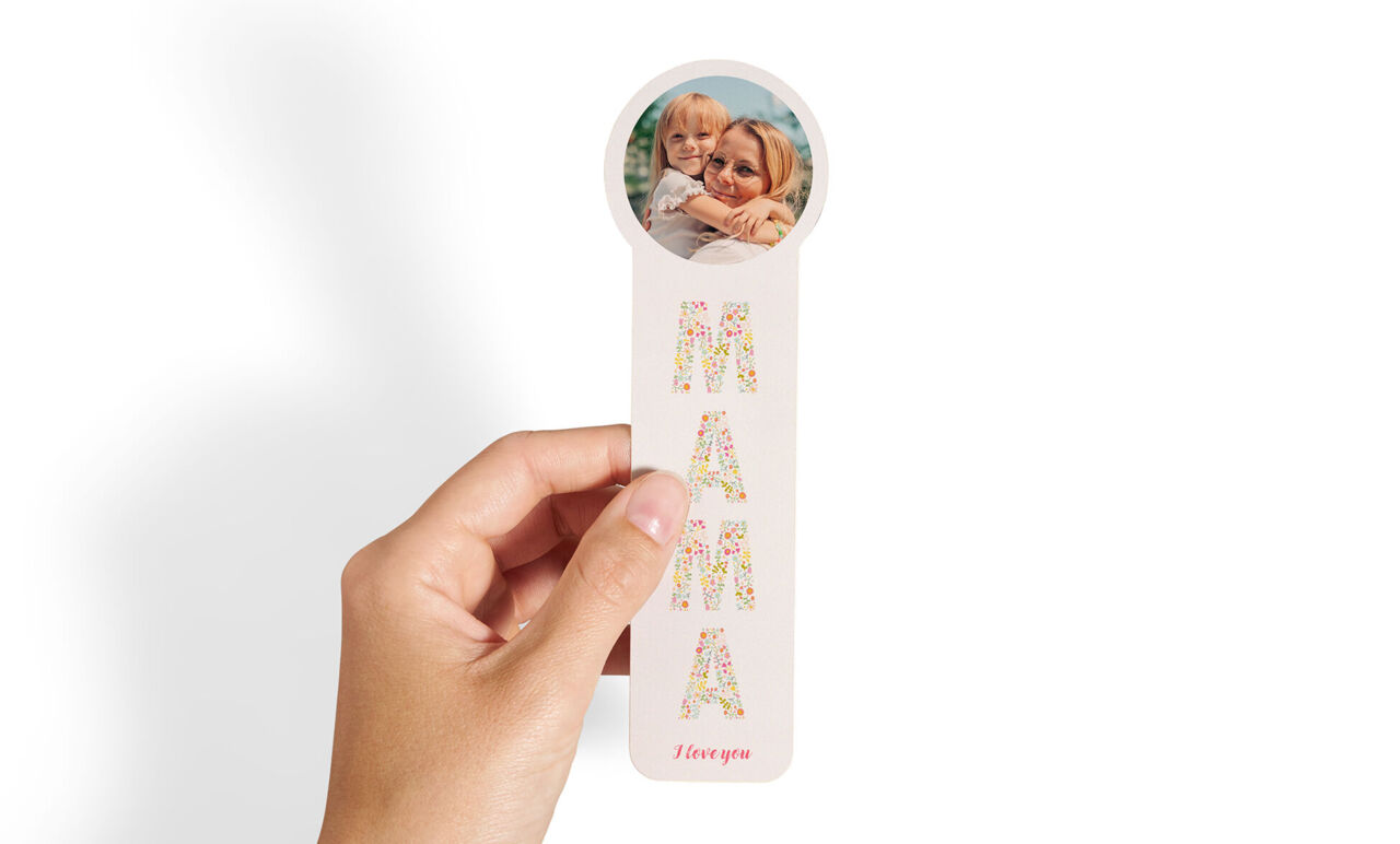 Eine Hand hält ein Lesezeichen mit Aufschrift "Mama" und einem Kinderfoto im runden Kopfteil