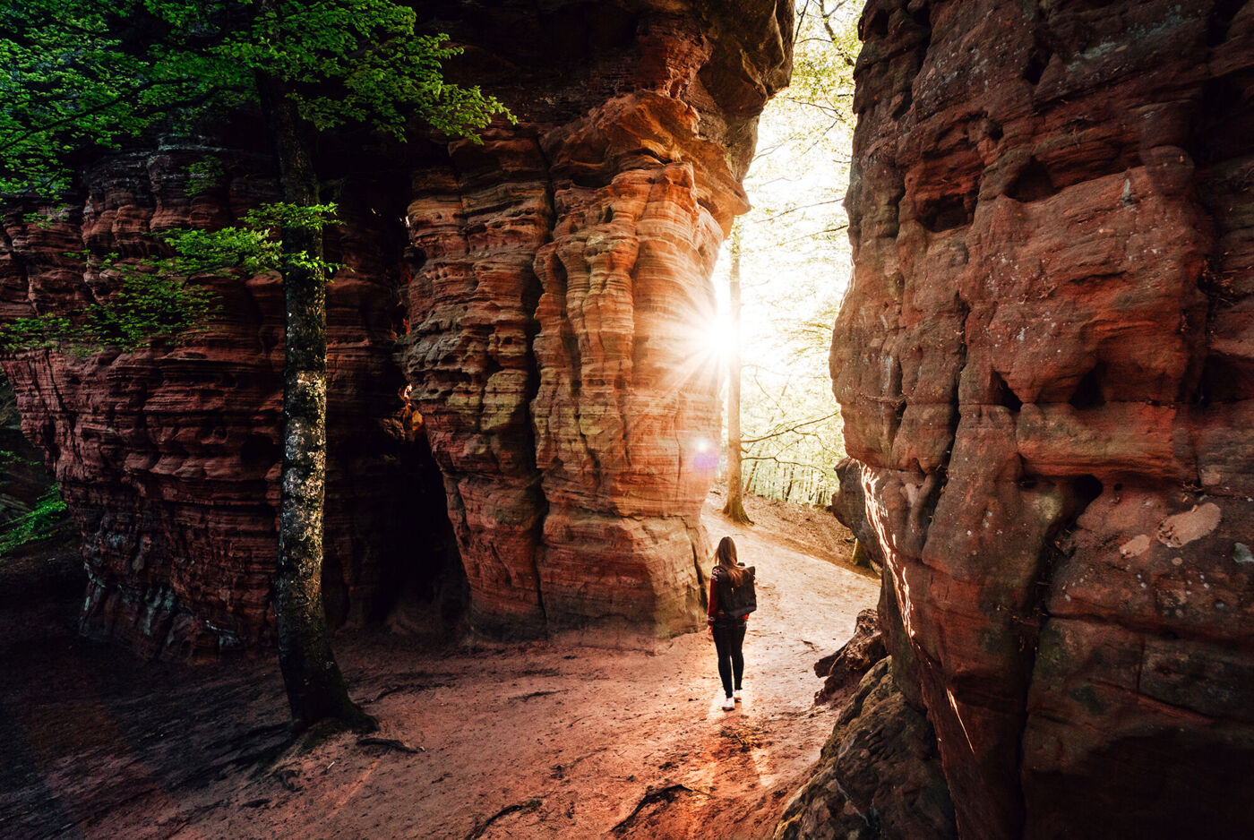 Eine Person, die zwischen zwei großen Felsen auf einem Weg spaziert. Im Hintergrund sieht man einen Sonnenuntergang.