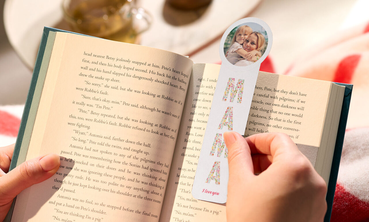 Eine Hand legt ein Lesezeichen in ein aufgeschlagenes Buch. Das Lesezeichen ist oben abgerundet, mit Aufschrift Mama und einem Kinderfoto