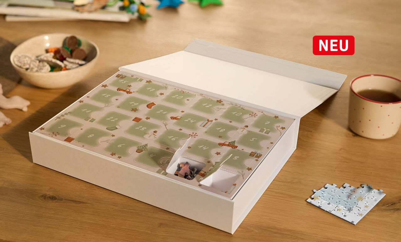 Ein Adventskalender liegt auf einem Tisch. Hinter geöffneten Türchen sind Puzzleteile zu erkennen, die mit eigenem Foto bedruckt sind