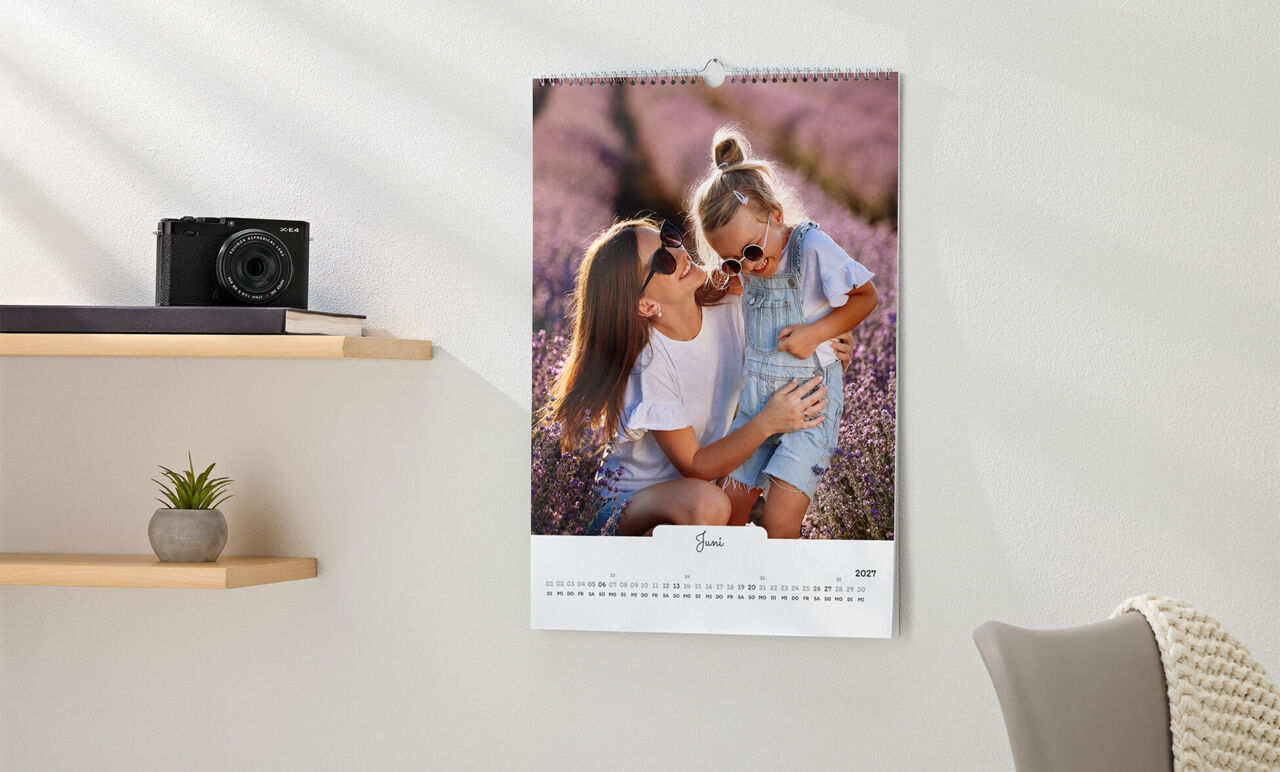Kalender im Hochformat mit weißem Design in handgeschriebenen Stil, mit Kinderfoto, hängt an der Wand neben einem Regal