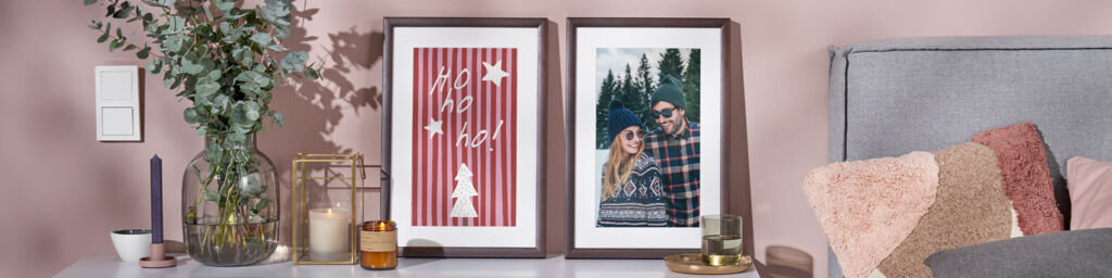 Zwei Poster im Rahmen stehen auf einer Kommode neben einem Sofa. Das linke zeigt ein Weihnachtsdesign, das rechte ein Paarfoto im Schnee