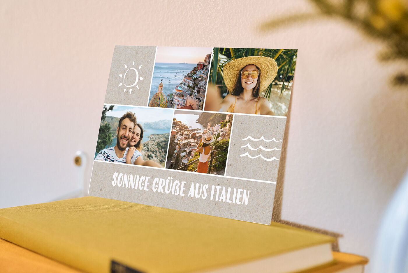 Eine Postkarte mit einer Collage aus Urlaubsfotos und dem Titel "sonnige Grüße aus Italien" steht auf einem Bücherstapel an einer Wand