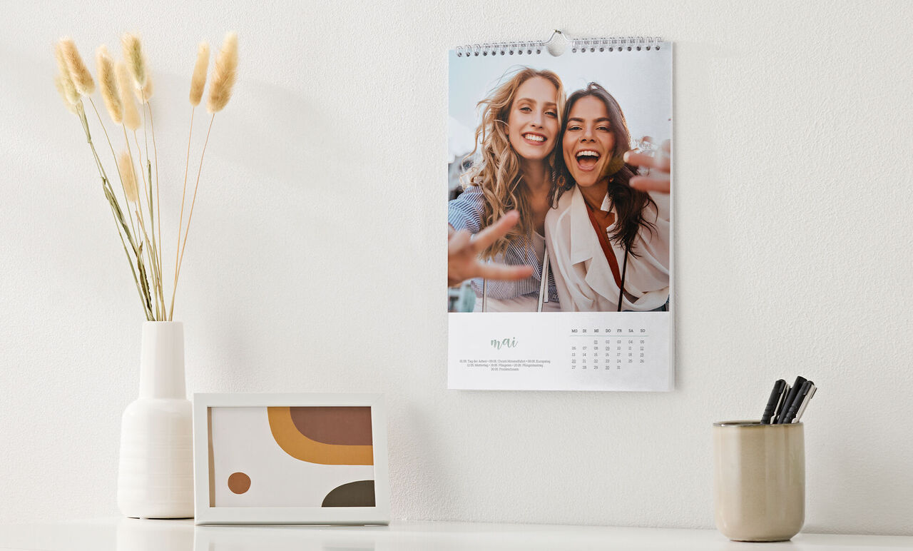 Kalender im Hochformat und weißem Design, mit Foto von Freundinnen, hängt über Sideboard